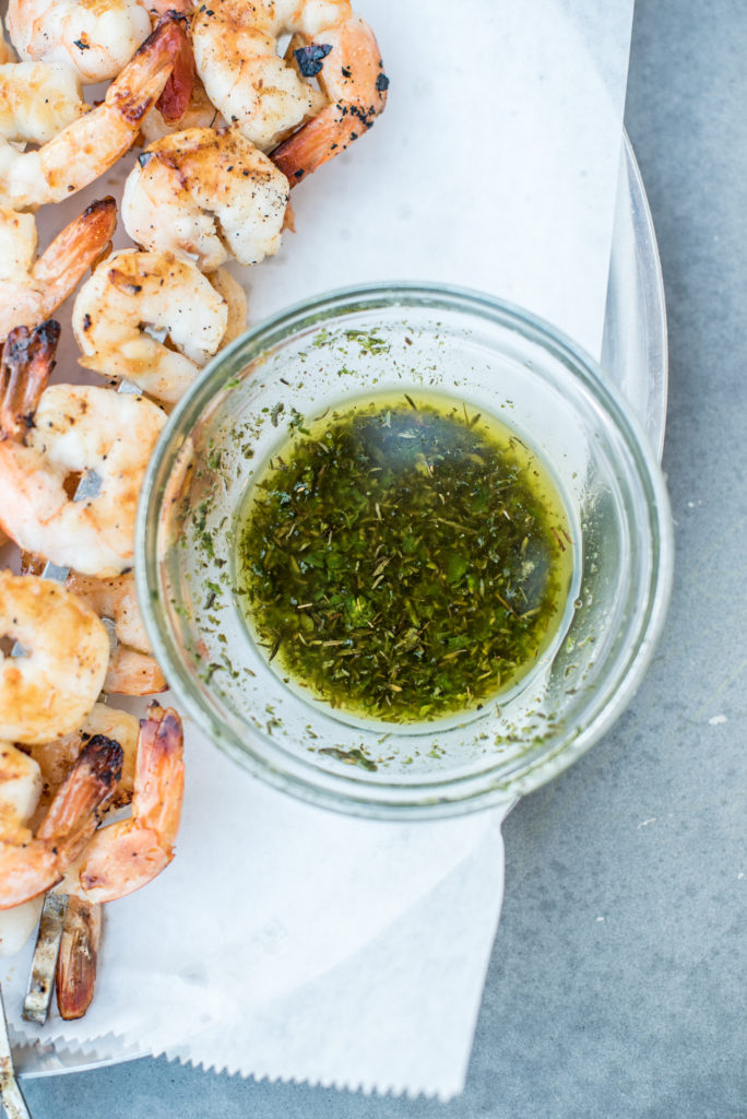 Chimichurri sauce in a glass dish next to cooked honey cajun shrimp skewers on a metal platter.
