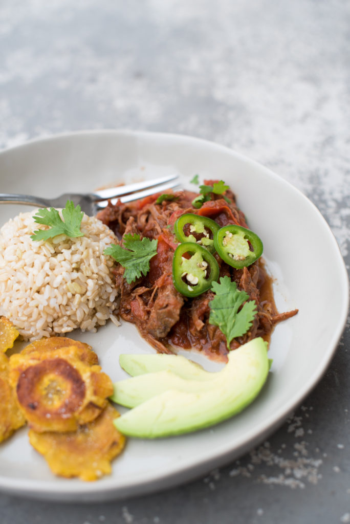 bowl with shredded Ropa Vieja, fried plantains, and a serving of rice on a gray table